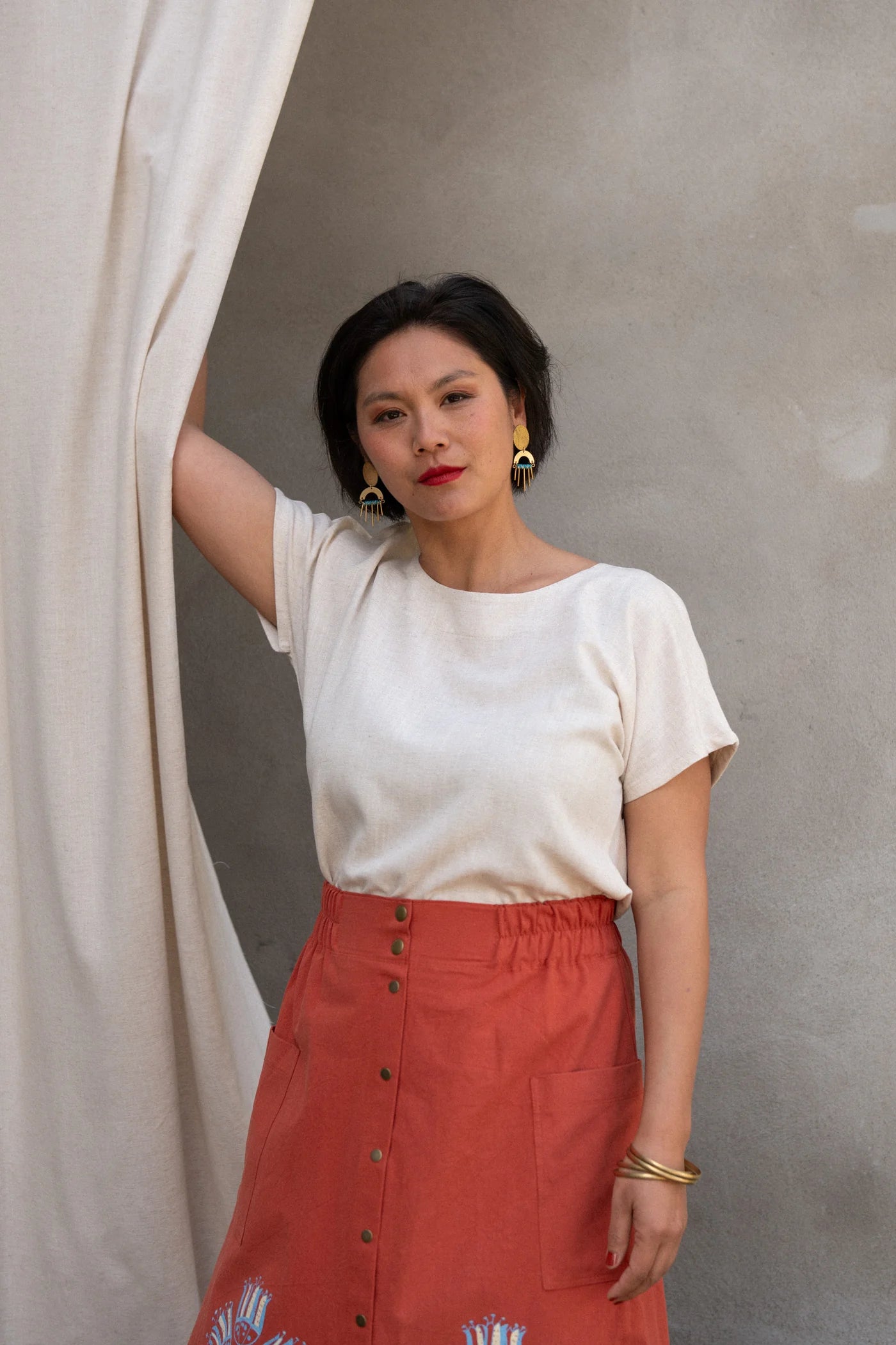  A woman wearing the Nymphea Screenprinted Skirt by Kazak in Red Pysanka with a cream top, she is standing outside in front of white curtain. 