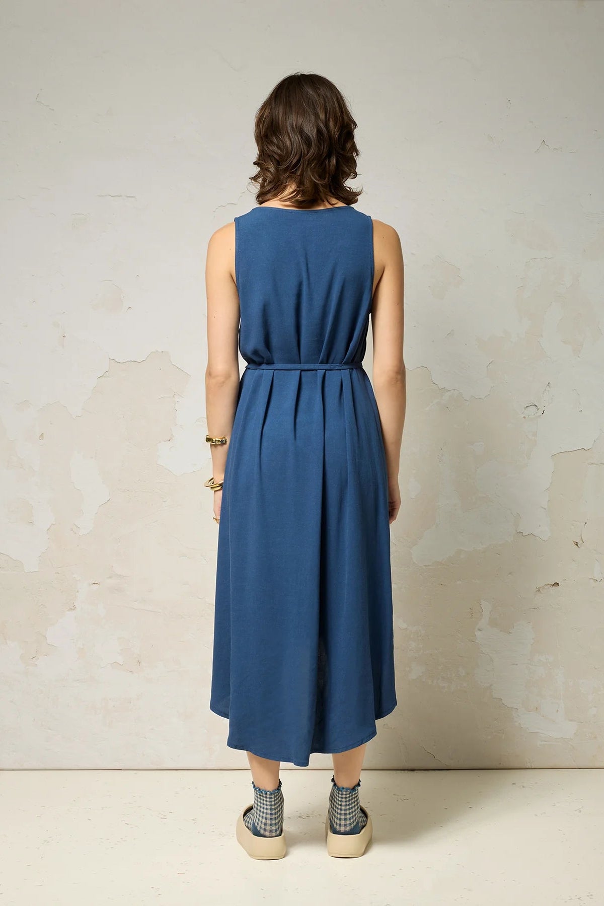 Back view of a woman wearing the Chaos Dress by Cokluch in Denim Blue, a long loose sleeveless dress with a V-neck, matching belt, and hi-low hemline. She is standing against a plain background