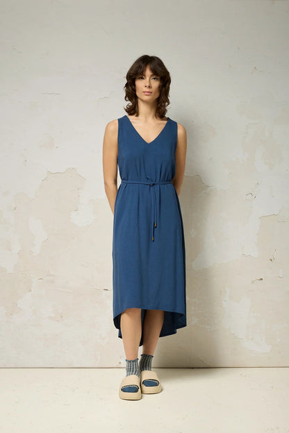 A woman wearing the Chaos Dress by Cokluch in Denim Blue, a long loose sleeveless dress with a V-neck, matching belt, and hi-low hemline. She is standing against a plain background