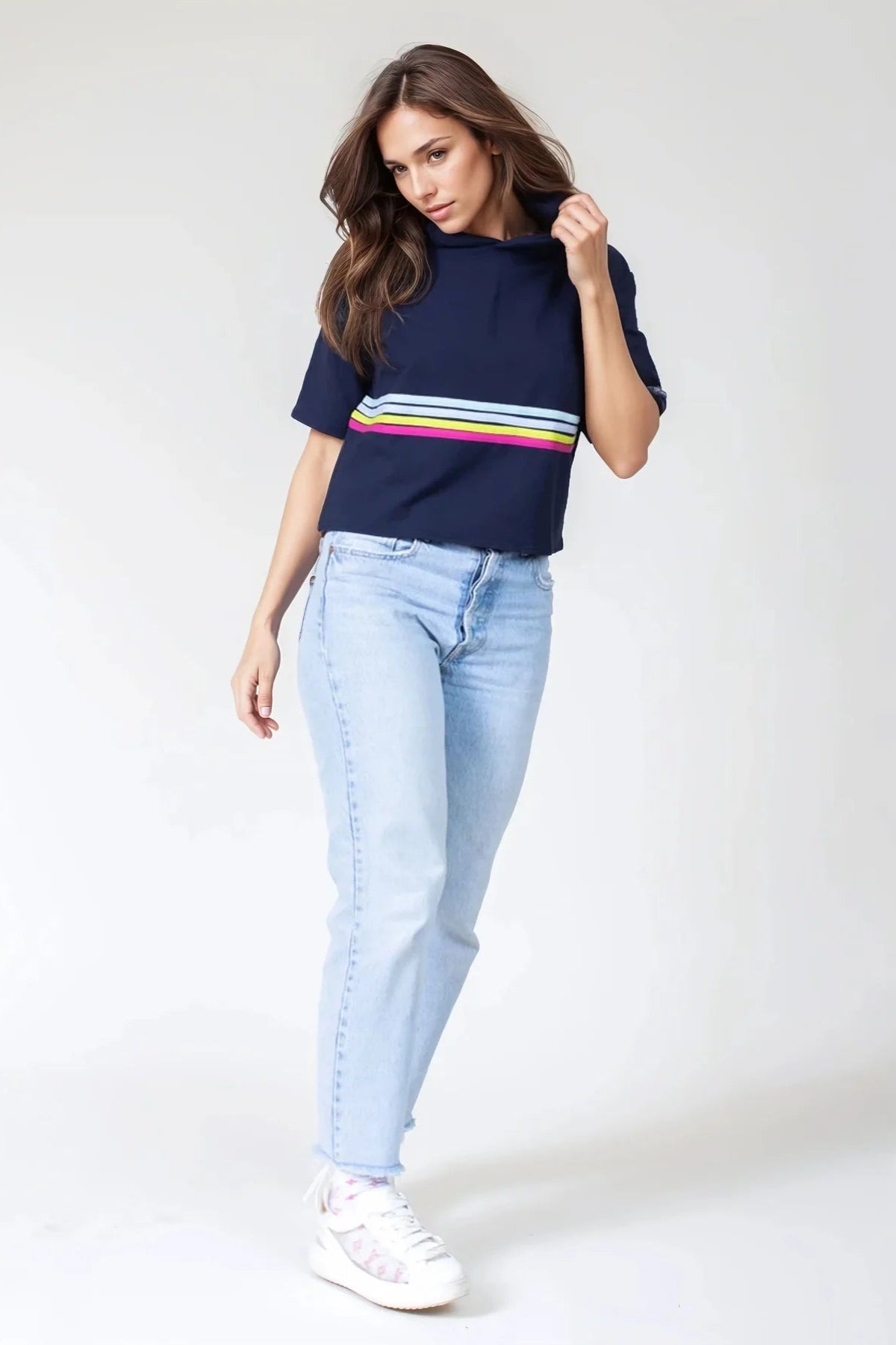A woman wearing the Olive+Splash Bamboo T-Shirt Hoodie in Striped navy,  featuring short sleeves, a cropped fit, and a splash of bright stripes across the midsection with a vertical gold zipper detail at the right hemline. She is wearing it with jeans and standing in front of a white background. 