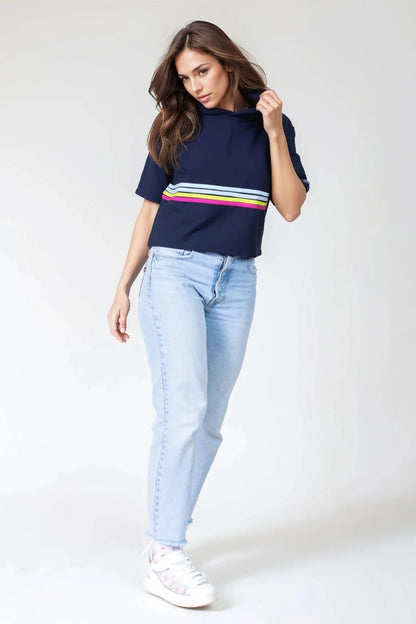 A woman wearing the Olive+Splash Bamboo T-Shirt Hoodie in Striped navy,  featuring short sleeves, a cropped fit, and a splash of bright stripes across the midsection with a vertical gold zipper detail at the right hemline. She is wearing it with jeans and standing in front of a white background. 