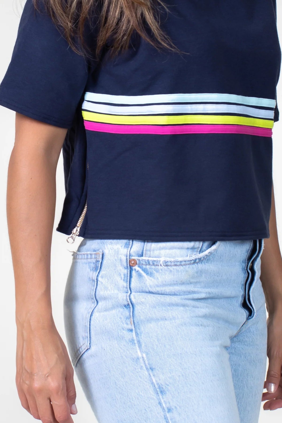 Close up detail of a woman wearing the Olive+Splash Bamboo T-Shirt Hoodie in Striped navy,  featuring short sleeves, a cropped fit, and a splash of bright stripes across the midsection with a vertical gold zipper detail at the right hemline. She is wearing it with jeans and standing in front of a white background. 