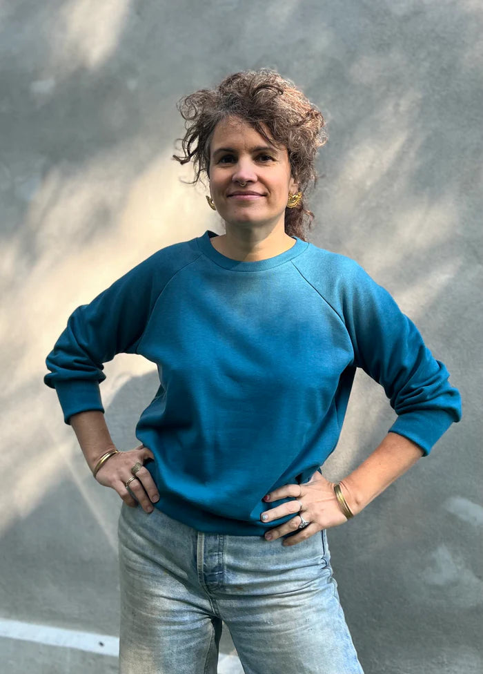 A woman wearing the Lipa Crewneck Sweatshirt by Kazak in Teal, featuring a round neck, 3/4 raglan sleeves, and a fitted band across the bottom. She is wearing it with jeans and is standing outside in front of a grey wall.