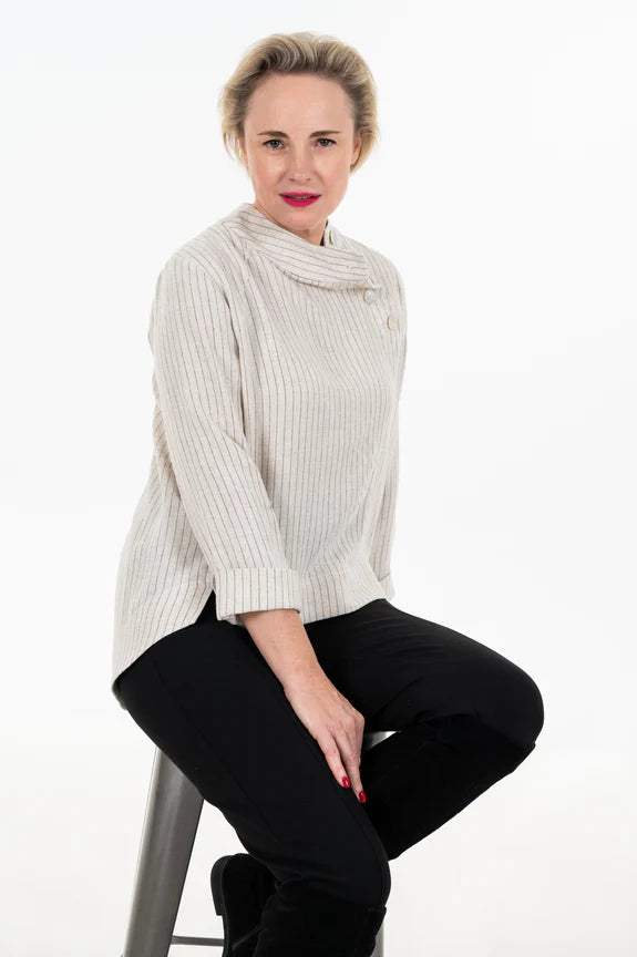 A woman wearing the Charlie Top by Julei Design in Natural Stripe with dark pants, seated on a stool in front of a white background 