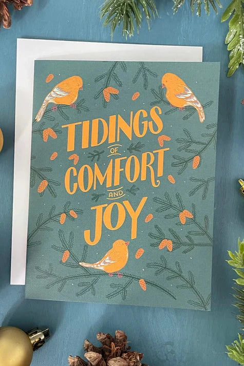 A Goldfinch card from a box set by Carabara Designs, featuring goldfinches and gold lettering that reads "Tidings of Comfort of Joy" on a teal background, is shown on a teal background surrounded by holiday decor. 