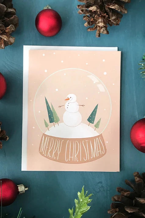 A card from a box set by Carabara Designs, featuring a snowman in a snow globe and the words "Merry Christmas" is shown against a teal background with holiday decor around it. 