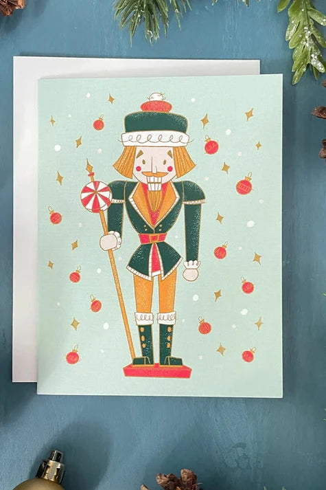 A card from a box set by Carabara Designs, featuring a Midcentury Nutcracker design on a pale blue background, is shown against a teal background with holiday decor around it. 