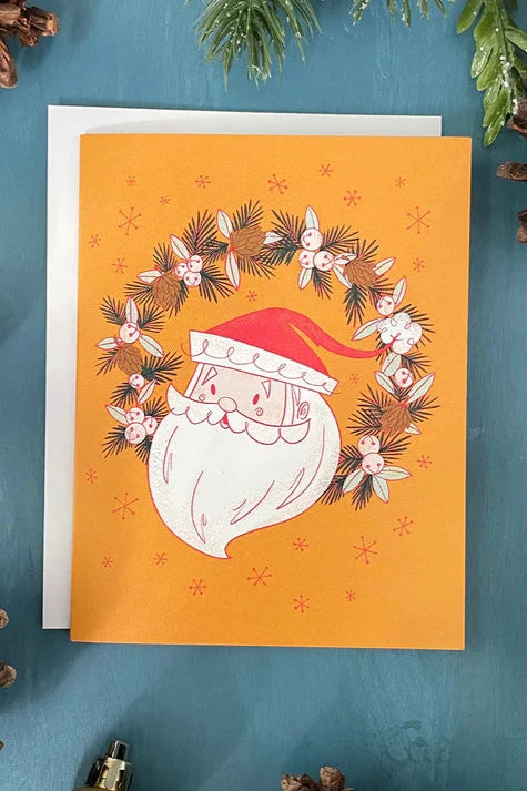 A card from a box set by Carabara Designs, featuring a Midcentury Santa design on a yellow background, is shown against a teal background with holiday decor around it. 