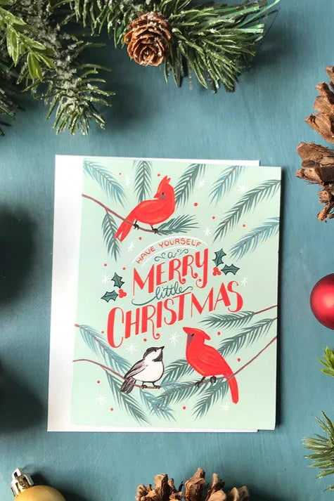 A card from the Carabara Design's Cardinal Christmas Card box set, featuring hand-drawn cardinals and greenery and reading "Have Yourself a Merry Little Christmas, is shown against a teal background with holiday decor around it. 