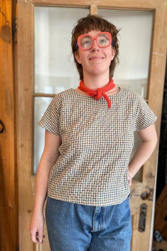 A woman wearing the Boronia Top by Kazak in Multicoloured Tiles with a round neck, short sleeves, slightly cropped and boxy fit, with a box pleat at the back yoke. She is wearing it with a red scarf and light jeans and standing in front of a wooden door. 