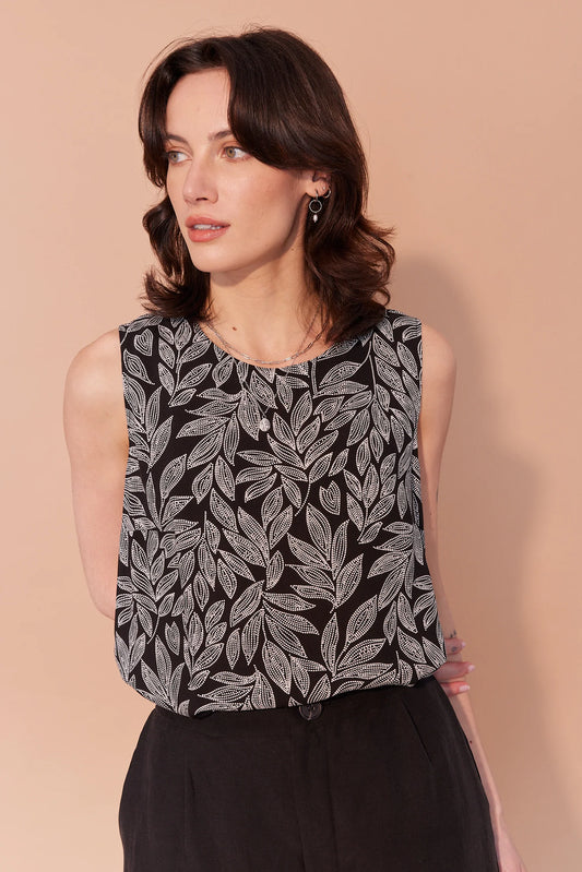 A woman wearing the Helene Cami from Cherry Bobin in Black and White Floral, with a round neck, wide straps, a loose flowing fit, and side slits. She is wearing it with black pants and is standing in front of a beige background. 