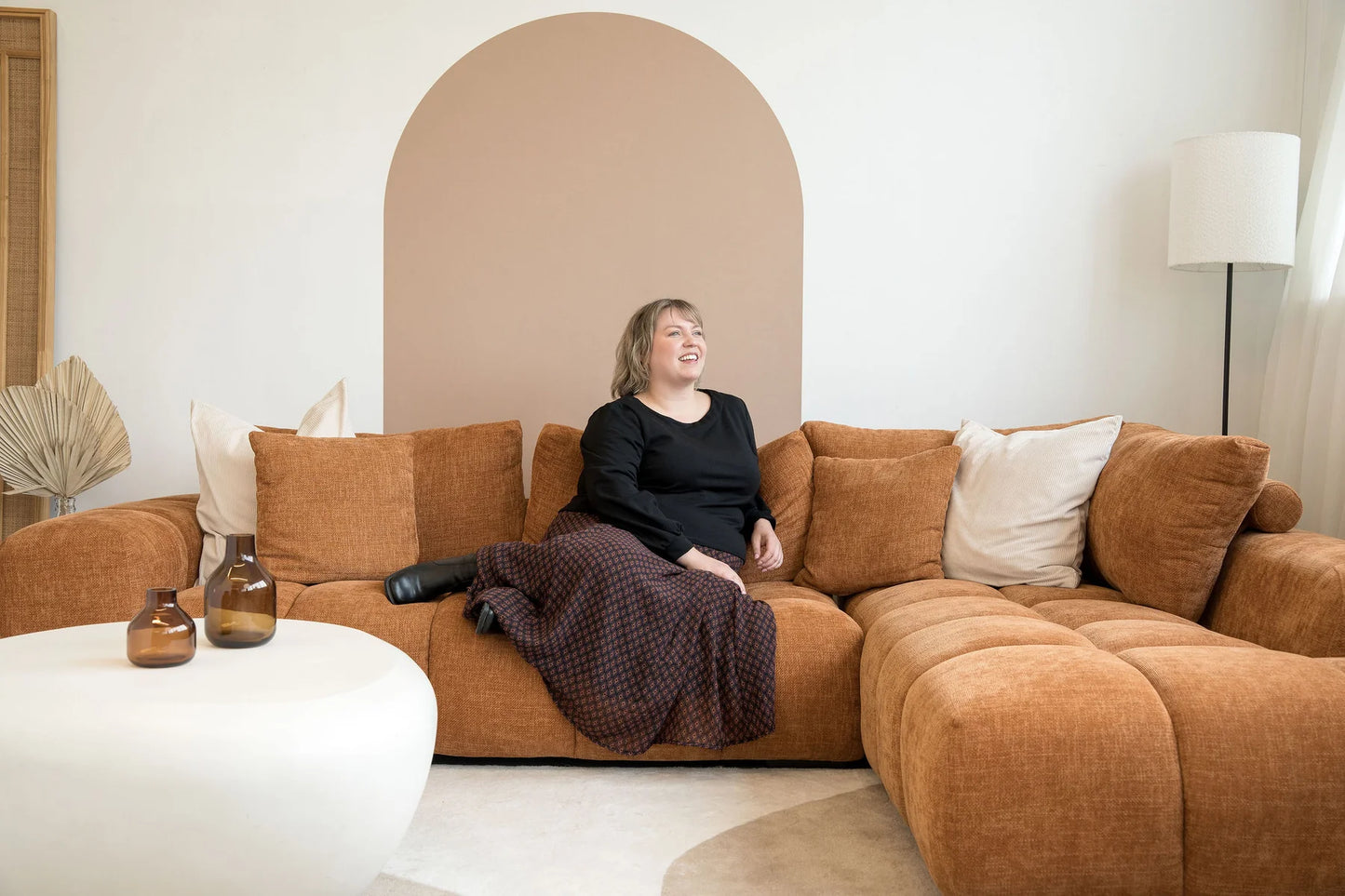 A woman wearing the Espresso Skirt by Cherry Bobin in Black and Brown print, a long and flowing chiffon skirt that's fully lined. She's wearing it with a black top and sitting on a brown sofa. 