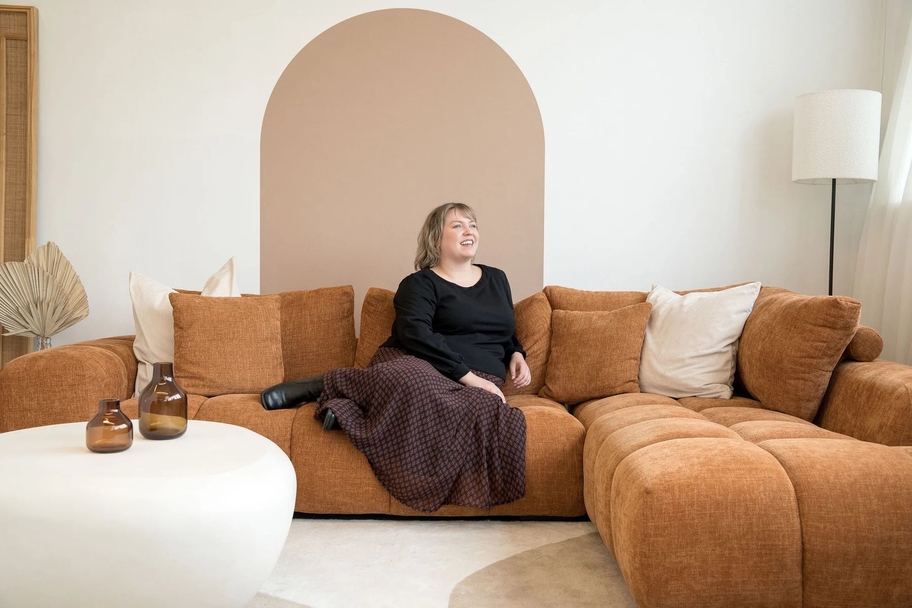 A woman wearing the Espresso Skirt by Cherry Bobin in Black and Brown print, a long and flowing chiffon skirt that's fully lined. She's wearing it with a black top and sitting on a brown sofa. 