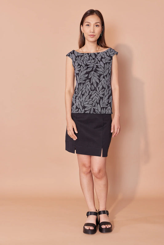 A woman wearing the Simone Cami by Cherry Bobin in Black and White Leaves, with a wide boat neck that just covers the shoulders, a fitted shape, and a hip length. She is wearing it with a black skirt and standing in front of a beige background. 