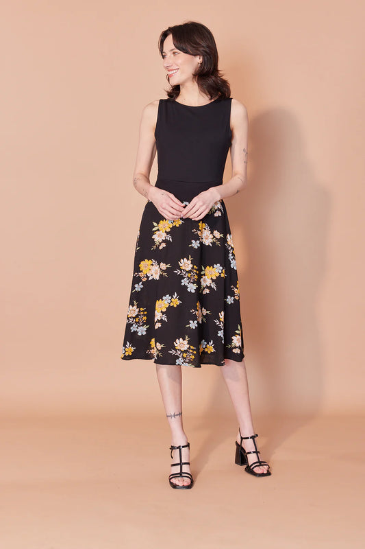 A woman wearing the Sofia Dress by Cherry Bobin in Orange Floral, featuring high-necked sleeveless bodice in black Tencel modal, with a flowing patterned skirt that hits below the knee. She is standing in front of a beige background. 