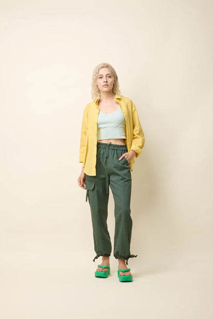 A woman wearing the Chila Shirt by Cokluch in Banana, a button down shirt with a blouse collar, rounded hem that falls below the hip, and long sleeves that can be cinched with a button and strap closure. She is wearing it with a lilac coloured crop-top and green pants and standing in front of a white background. 