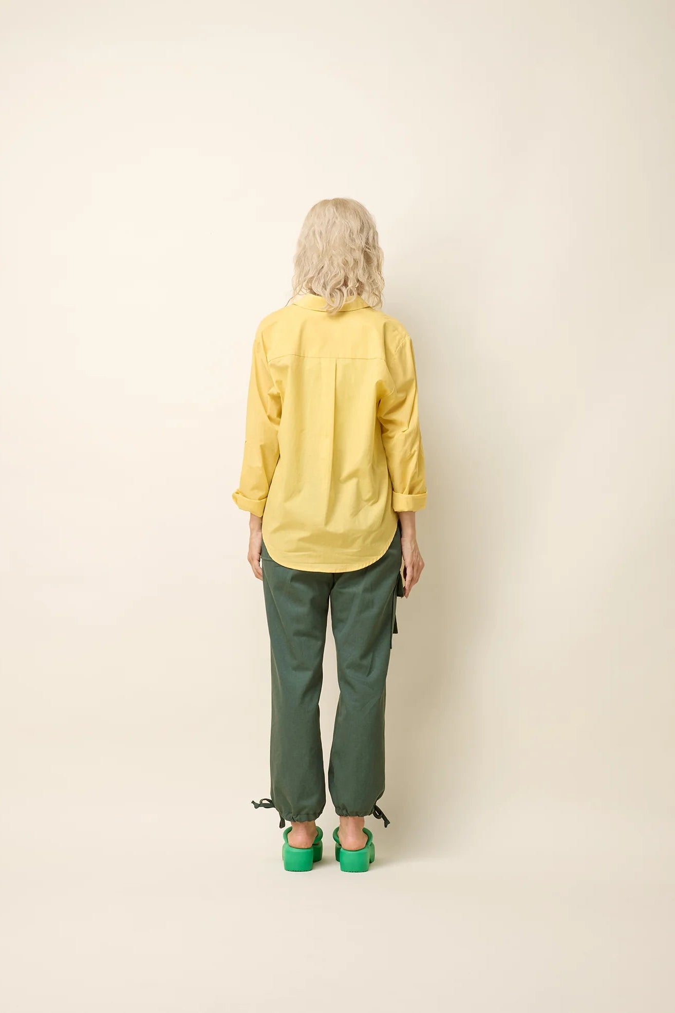 Back view of a woman wearing the Chila Shirt by Cokluch in Banana, a button down shirt with a blouse collar, rounded hem that falls below the hip, and long sleeves that can be cinched with a button and strap closure. She is wearing it with a lilac coloured crop-top and green pants and standing in front of a white background. 