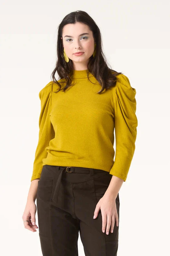 A woman wearing the Nenuphar Top by Cokluch in Avocado, featuring a high collar and 3/4 length puffed sleeves, with the Belief Pants in Pine. She's standing in front of a white background. 