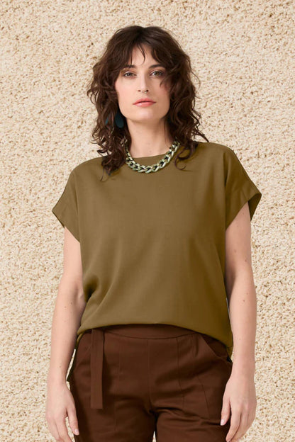 Semi close-up of a woman wearing the Ombre Top by Cokluch in Oregano, featuring a round neck, short dropped sleeves and a loose fit, with brown pants,  standing in front of a beige wall 