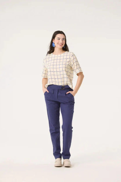 A woman wearing the Saules Pants by Cokluch in Twilight with a fly closure, an attached D-ring belt, a button and tab detail, side pockets, and an ankle length, made from OEKO-TEX certified fabric. She is wearing them with the Sideral Top in Cream Plaid and standing in front of a white background. 