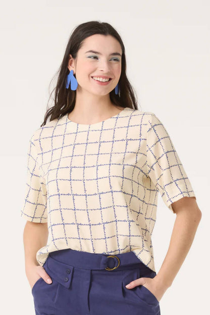 A semi-close up of a woman wearing  the Sideral Top by Cokluch in Cream Plaid with blue pants, standing in front of a white background 