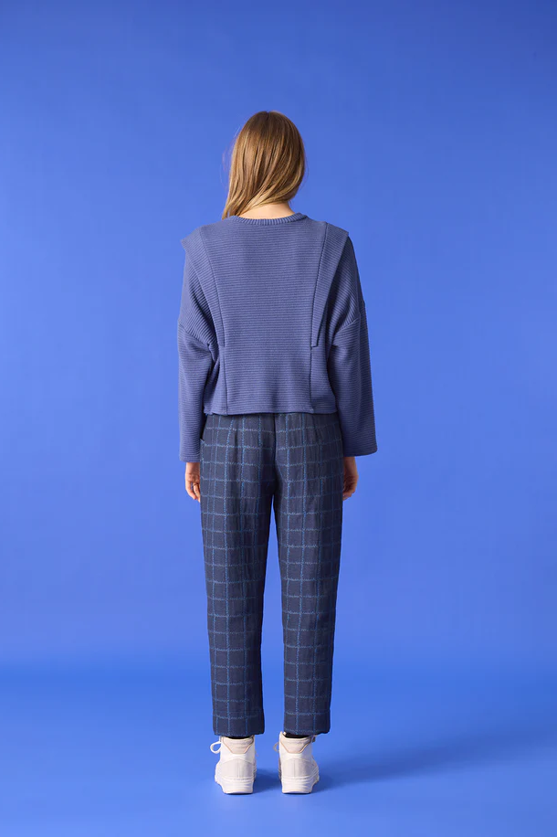  A woman wearing the Vega Pants by Cokluch in Denim, featuring an exclusive checked jacquard fabric and slim ankle-length legs and a pull on waist. She is wearing a Blue top and standing in front of a blue background. 