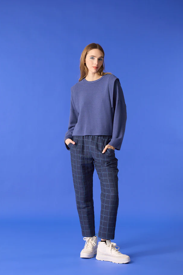  A woman wearing the Vega Pants by Cokluch in Denim, featuring an exclusive checked jacquard fabric and slim ankle-length legs and a pull on waist. She is wearing a Blue top and standing in front of a blue background. 