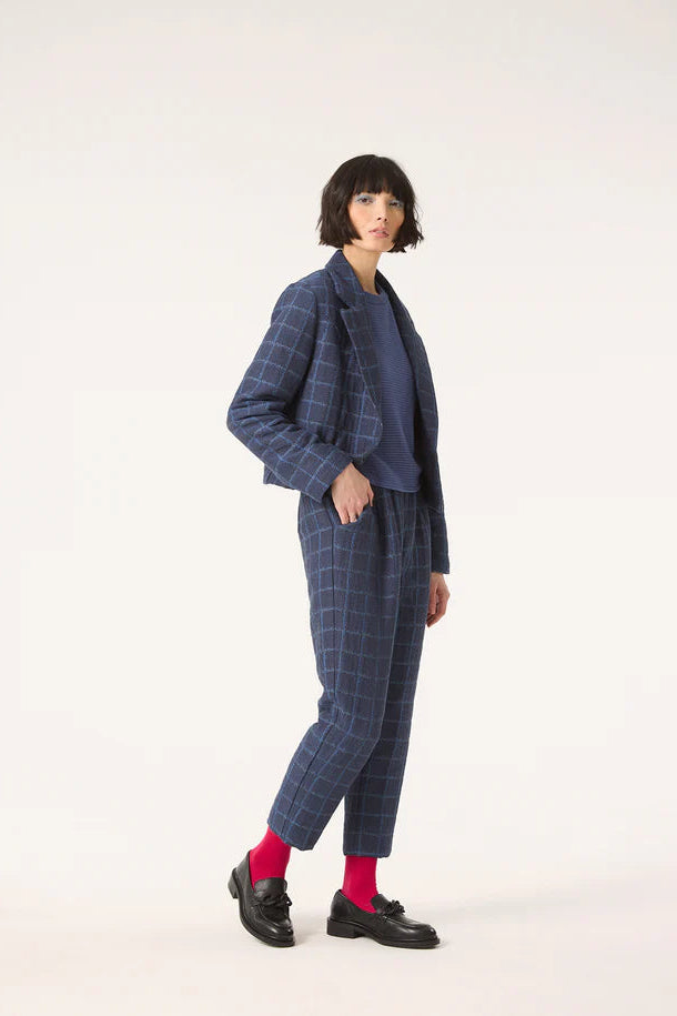 A woman wearing the Unreal Jacket by Cokluch in Denim, featuring a subtle plaid pattern, an open tailored collar, a short and slightly loose fit, and a two button closure. She is wearing with with a blue top and the matching Vega pants and is standing in front of a white background. 