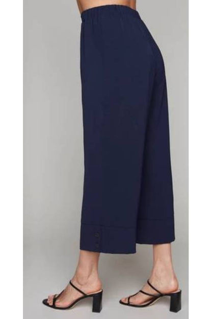 A side waist-down view of a woman wearing the Opal Pant by Compli K in Navy, with a high waist, wide cropped legs, and a button detail on the cuffs. She's standing in front of a grey background. 