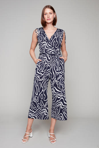 A woman wearing the Orville Jumpsuit by Compli K in a Navy and White Pattern, a sleeveless jumpsuit with ties at the shoulders, a V-neck, a faux-wrap waist, and wide cropped legs. She is standing in front of a grey background. 