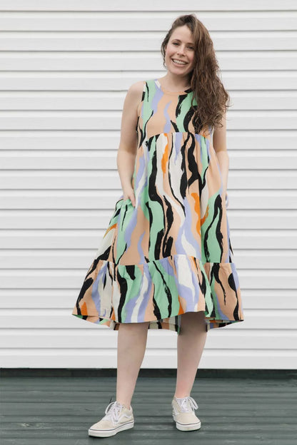 A woman wearing the Rose Dress by Copious in Mint/Peach abstract print, a sleeveless dress with an empire waist, tiered mid-calf length skirt, and pockets. She is standing against a white wall. 