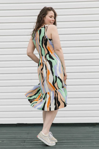 Side view of a woman wearing the Rose Dress by Copious in Mint/Peach abstract print, a sleeveless dress with an empire waist, tiered mid-calf length skirt, and pockets. She is standing against a white wall. 