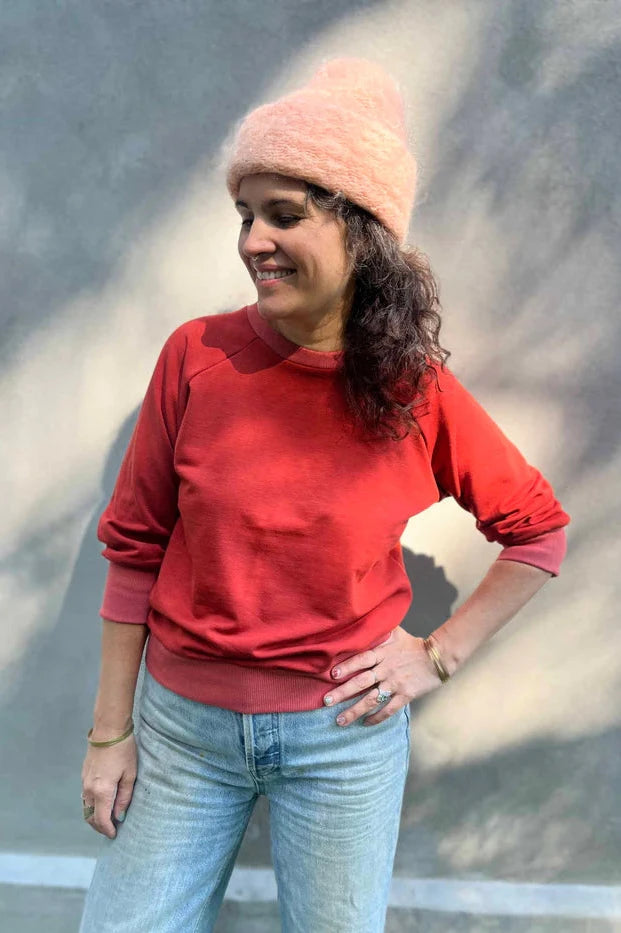 A woman wearing the Lipa Crewneck Sweatshirt by Kazak in Brick, featuring a round neck, 3/4 raglan sleeves, and a fitted band across the bottom. She is wearing it with jeans and a pink tuque and is standing outside in front of a grey wall. 