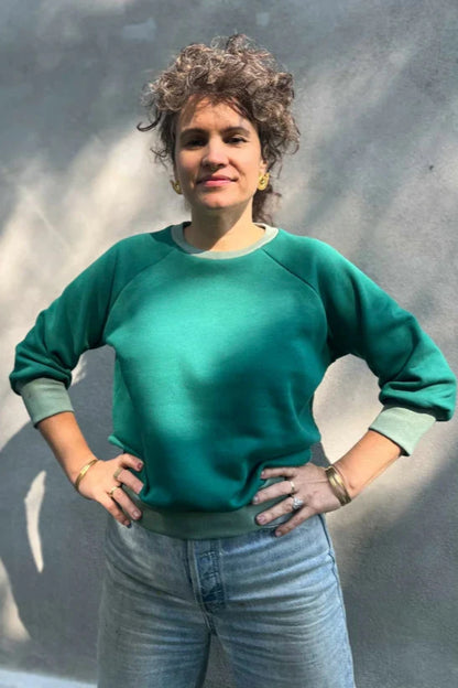 A woman wearing the Lipa Crewneck Sweatshirt by Kazak in Cedar and Sage, featuring a round neck, 3/4 raglan sleeves, and a fitted band across the bottom. She is wearing it with jeans and is standing outside in front of a grey wall.
