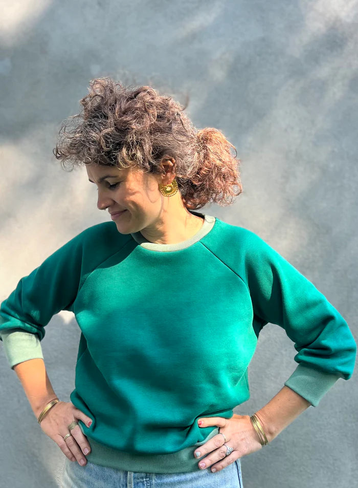 A woman wearing the Lipa Crewneck Sweatshirt by Kazak in Cedar and Sage, featuring a round neck, 3/4 raglan sleeves, and a fitted band across the bottom. She is wearing it with jeans and is standing outside in front of a grey wall.