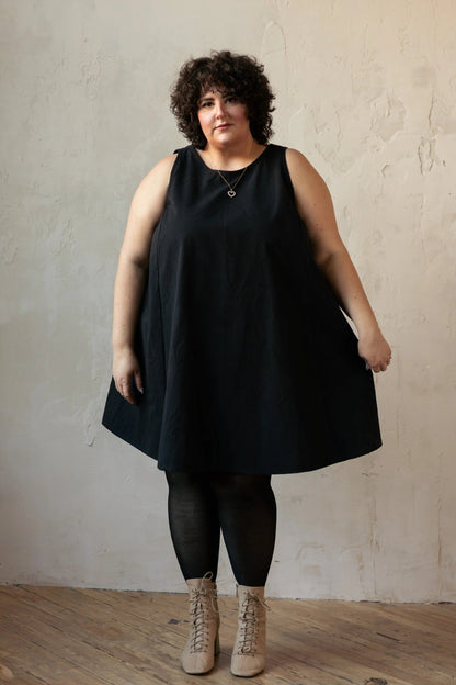 A woman wearing the Caro Dress by Lights of All in Black, a sleeveless knee-length trapeze dress with a round neck at the front and a V at the back. She is wearing it with black leggings and standing against a beige wall. 