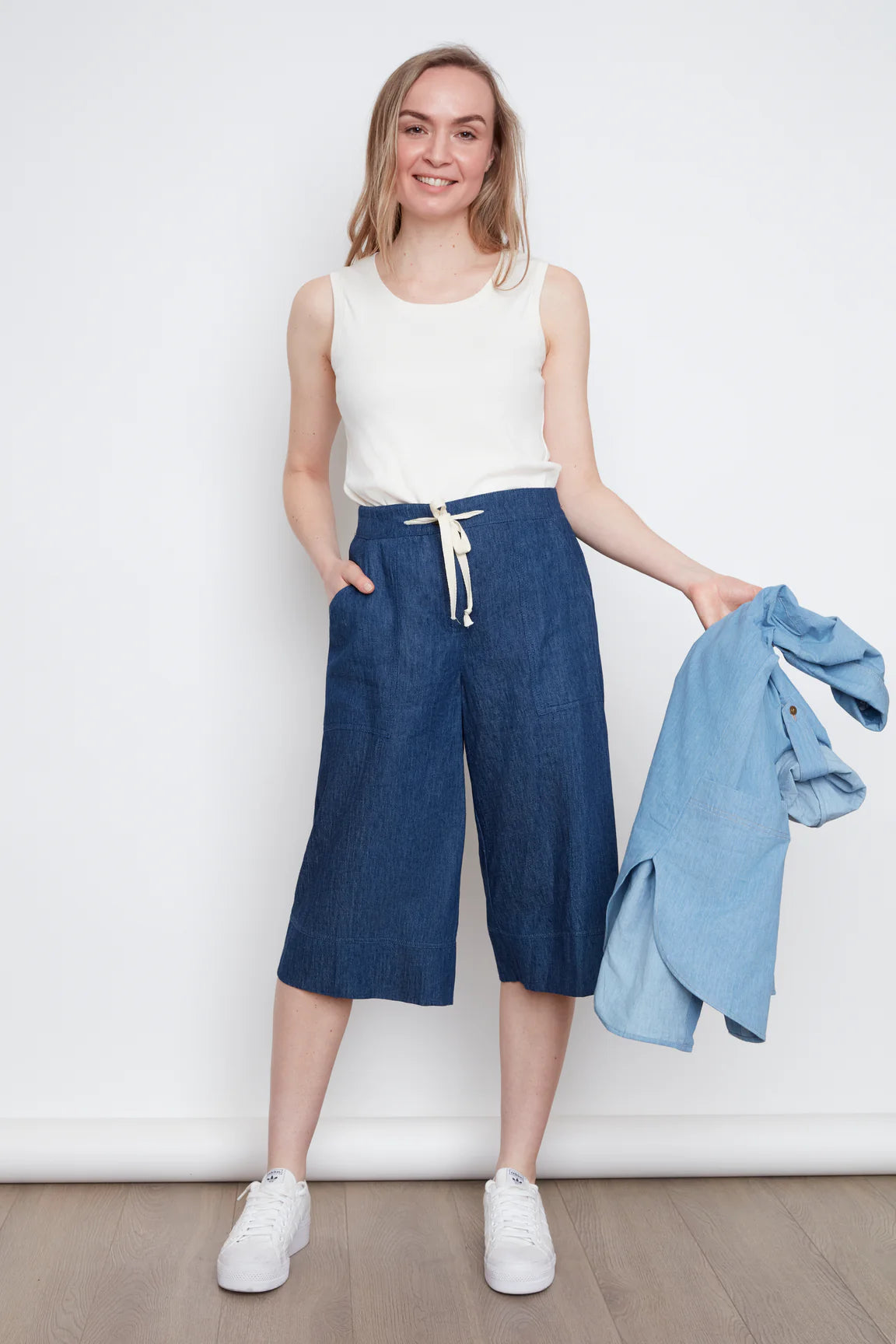 A woman wearing the Brooke Capri by Julei, wide-legged denim capris with a white drawstring and elastic at the back waist. She is wearing them with a white tank and holding a denim shirt while standing against a white wall.