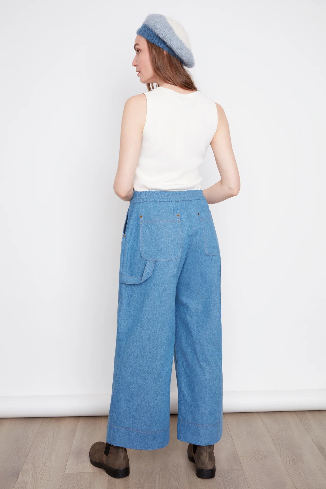 Back view of a woman wearing the Brooke Wide Leg Denim Pants by Julei in light denimm, with large patch pockets, a zipper, elastic at the back waist, and a carpenter loop. She is wearing them with a white top and standing in front of a white background. 