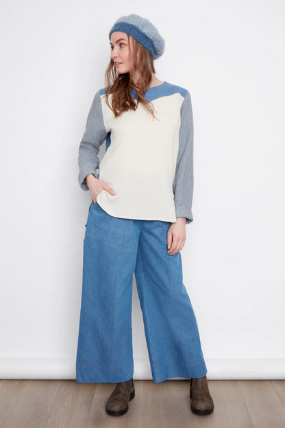 A woman wearing the Brooke Wide Leg Denim Pants by Julei in light denimm, with large patch pockets, a zipper, elastic at the back waist, and a carpenter loop. She is wearing them with a white and grey top and standing in front of a white background. 