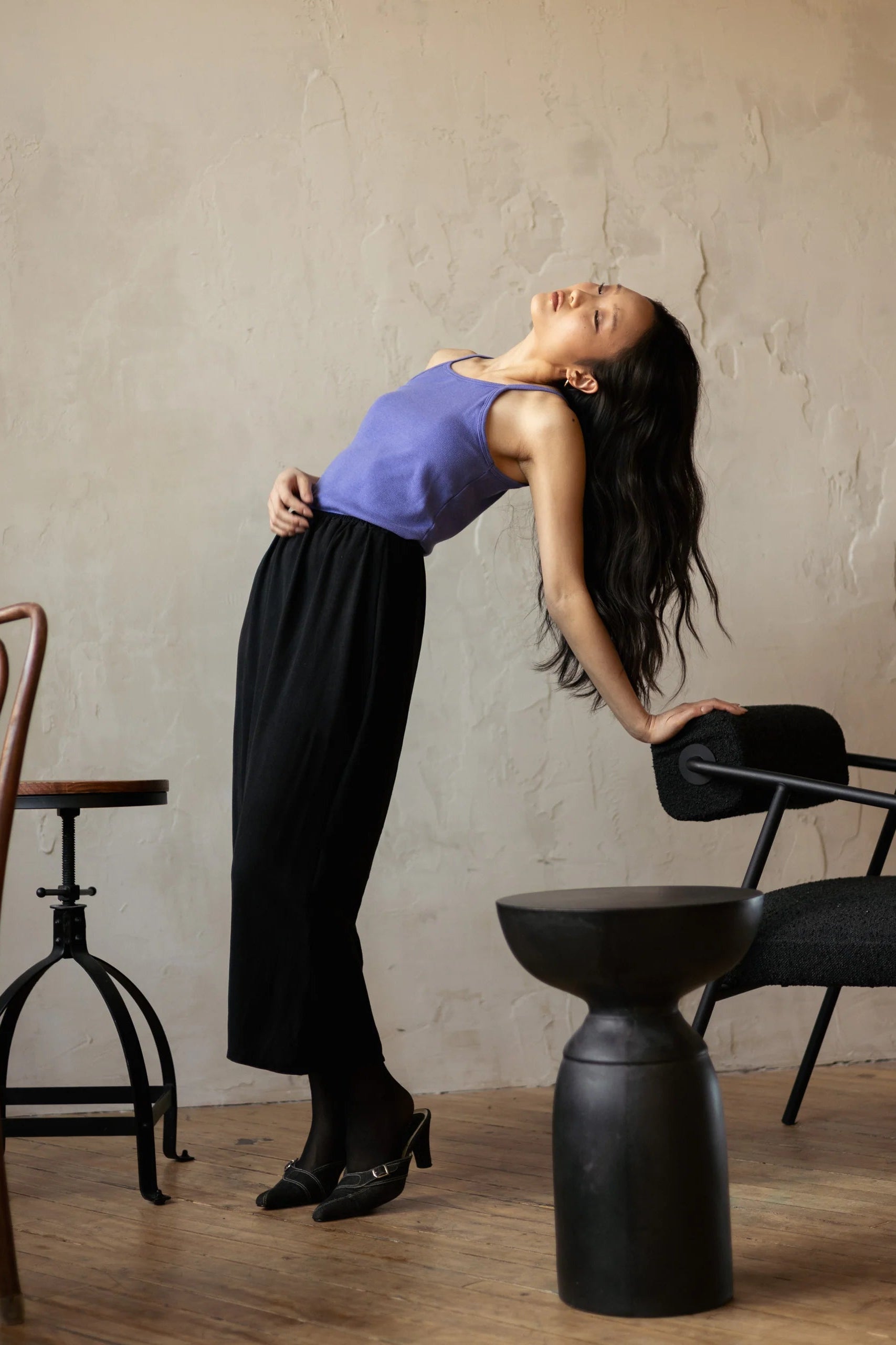 A woman wearing the Simone Skirt by Lights of All in Black, a sleek calf-length skirt with a flared hem and an elastic waist. She is wearing it with a purple top and is leaning back against a chair in a room with furniture. 