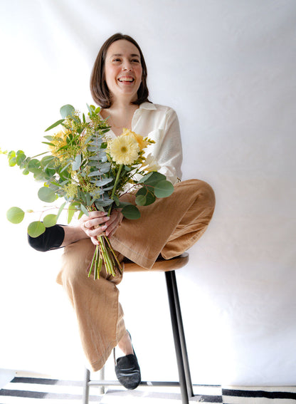 Woman wearing the Ania Blouse by Cokluch in Off-White, holding a bouquet of flowers sitting on a stool against a white wall