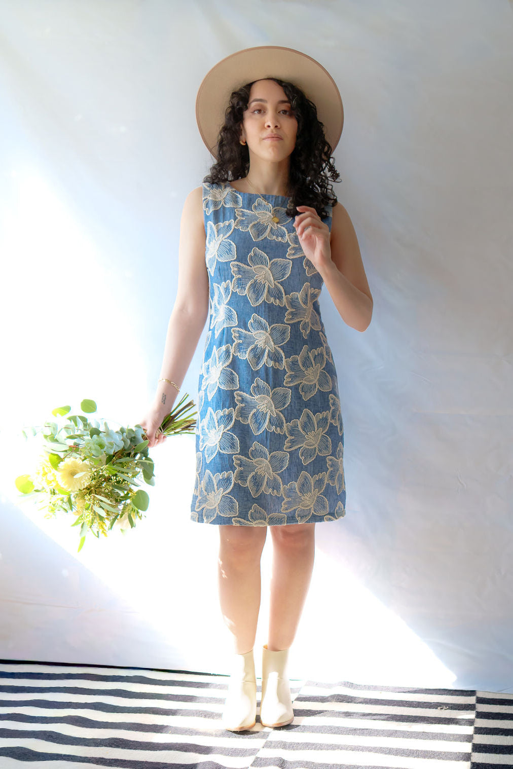 A woman wearing the Rose Dress by Dinh Ba, a sheath dress in pale blue with embroidered white flowers.  She is holding flowers and a tan hat and is  standing in front of a white background