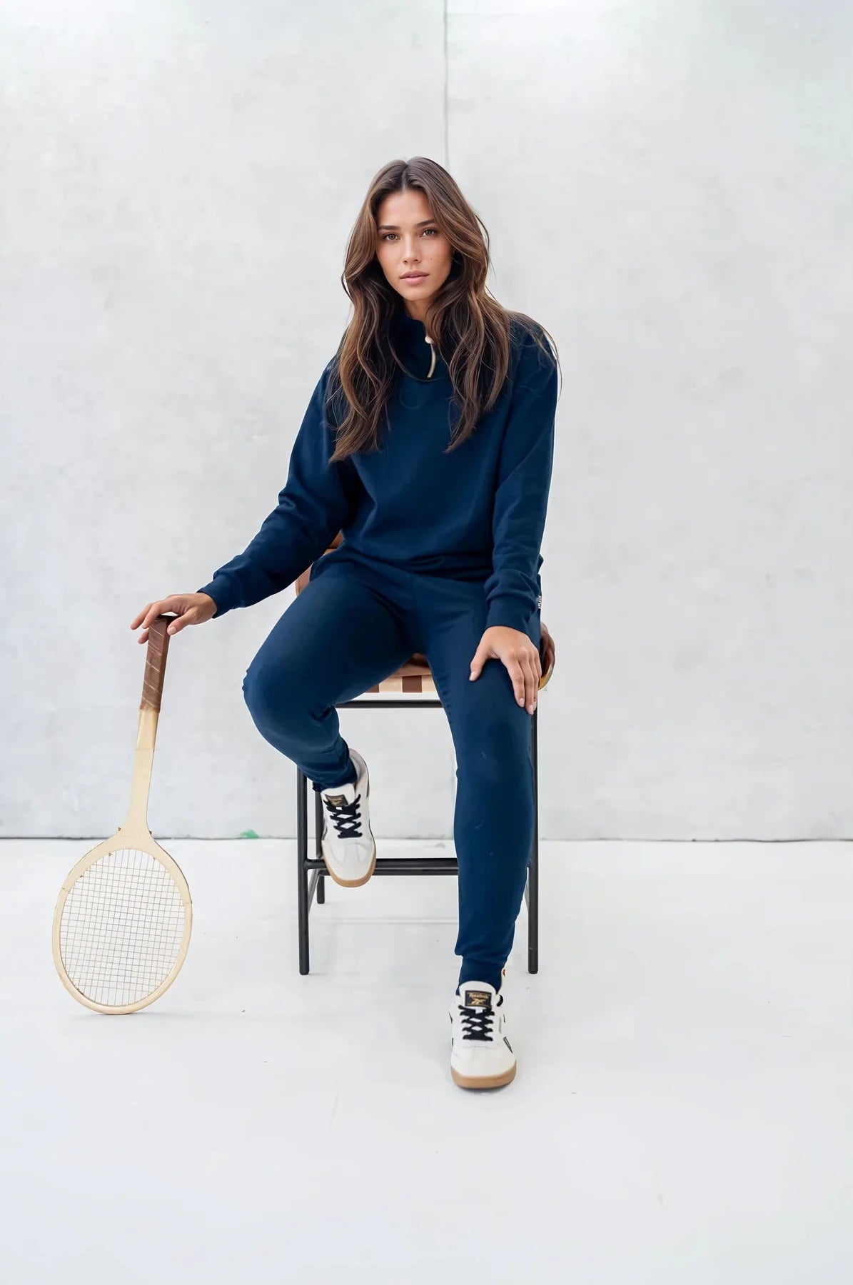 A woman wearing the 3/4 Zip Pullover by Olive+Splash in Navy, a long sleeved bamboo sweatshirt with a 3/4 length gold zipper. She is wearing it with matching pants and sitting on a chair in front of a white background. 
