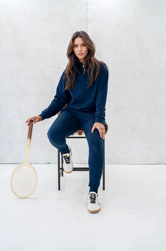 A woman wearing the 3/4 Zip Pullover by Olive+Splash in Navy, a long sleeved bamboo sweatshirt with a 3/4 length gold zipper. She is wearing it with matching pants and sitting on a chair in front of a white background. 