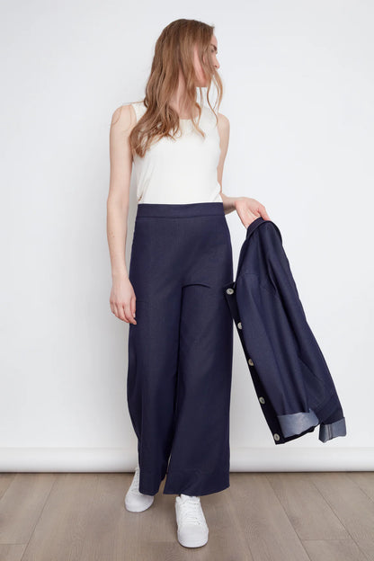 A woman wearing the Brooke Knit Trousers by Julei in Navy Knit, wide-leg pants with a pull-on waist, front slant pockets, and back patch pockets. She is wearing them with a white tank and holding a navy jacket against a white background.