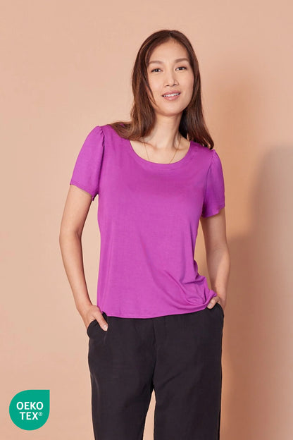 A woman wearing the Eugenie T-shirt by Cherry Bobin in Fuchsia, with a scoop neck, gathers at the shoulders, and a relaxed fit. She is wearing it with black pants and standing in front of a beige background. 