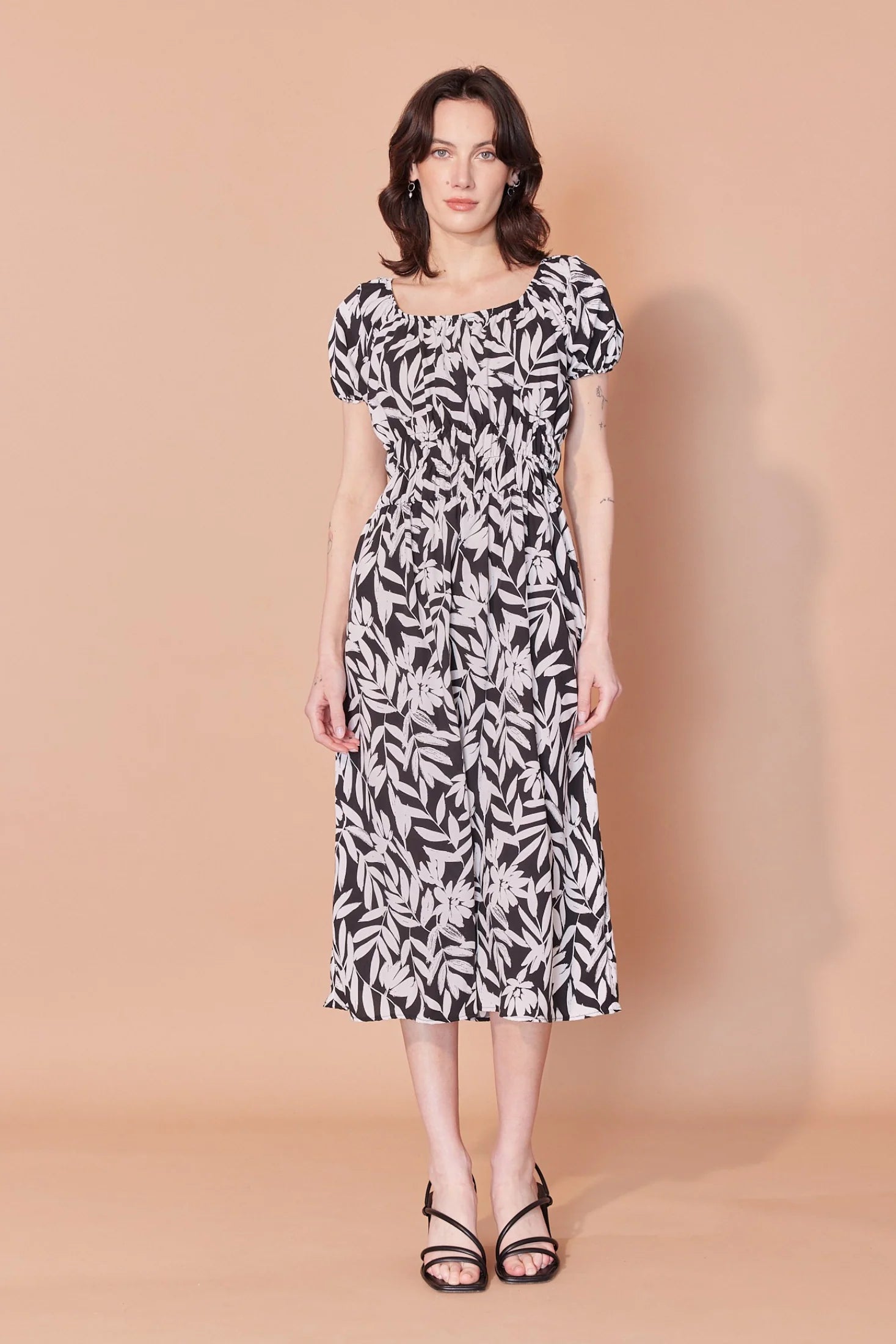 A woman wearing the Victoria Dress by Cherry Bobin in Black and Ivory Leaves print, featuring a scooped peasant neckline, short gathered sleeves, an empire waist with ruching and elastic, and a midi-length skirt. She is standing in front of a beige background. 