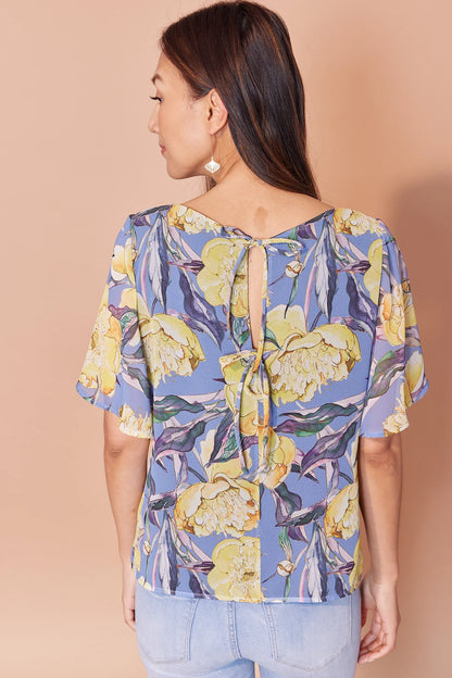 Back waist-up view of woman wearing the Anastasia Top by Cherry Bobin in Yellow Peony print, eaturing a v-neck, full elbow-length sleeves, and an open back with tie closures. She is wearing it with jeans and standing in front of a beige background. 