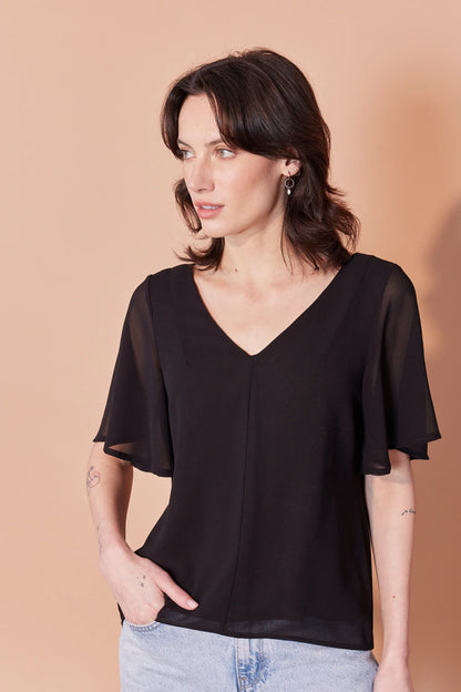 Waist-up view of a woman wearing the Anastasia Top by Cherry Bobin in Black, featuring a v-neck, full elbow-length sleeves, and an open back with tie closures. She is wearing it with jeans and standing in front of a beige background. 