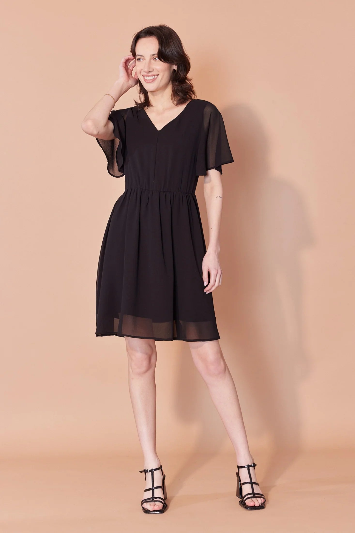 A woman wearing the Sisi Dress by Cherry Bobin in Black, a chiffon dress with a v-neck front and back, short full sleeves, a gathered waist, an open back with tie closures, and an A-line skirt that falls above the knee. She is standing in front of a beige background. 